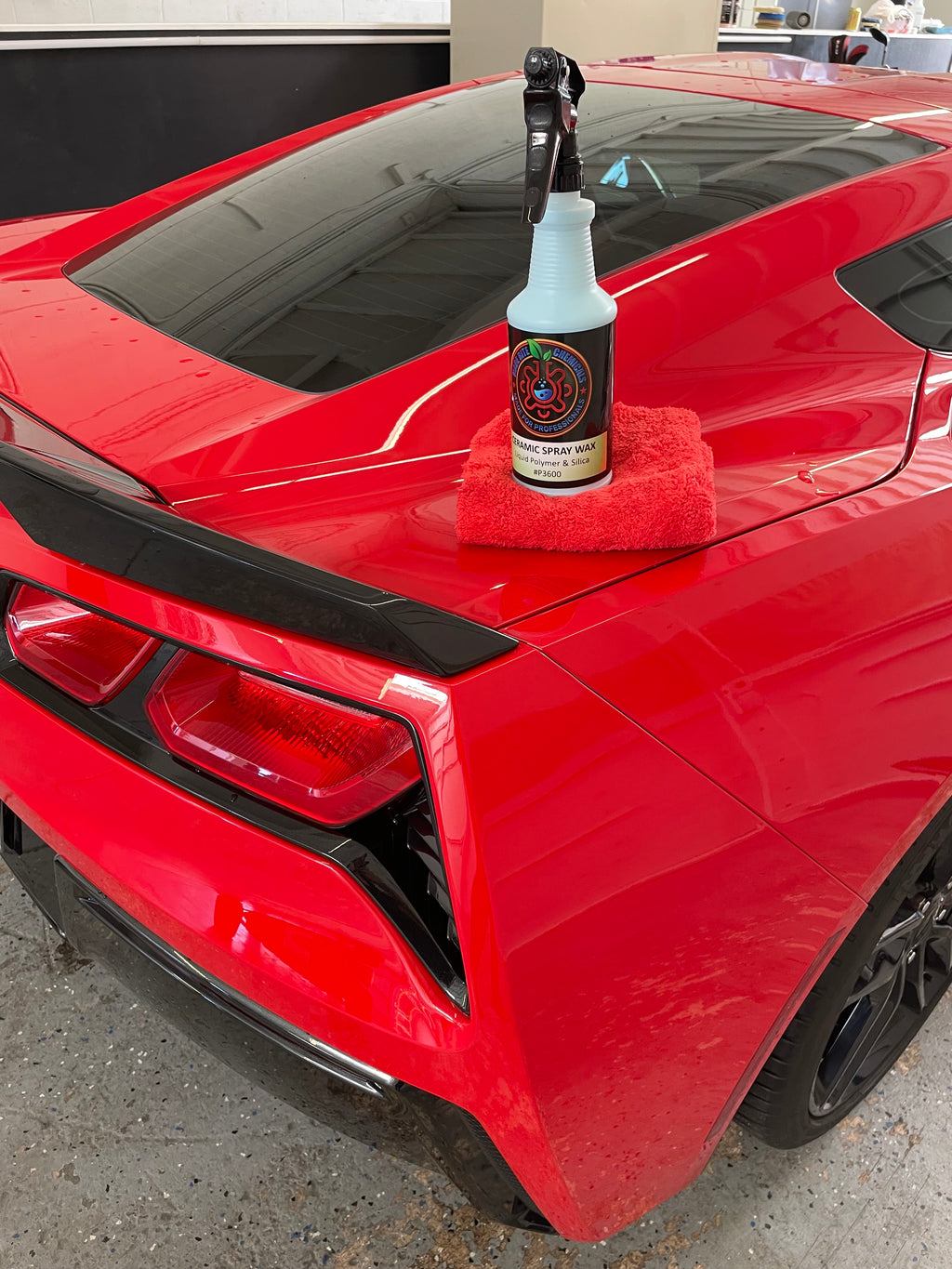 Red car with a bottle of wax attached to its rear, on a concrete surface.